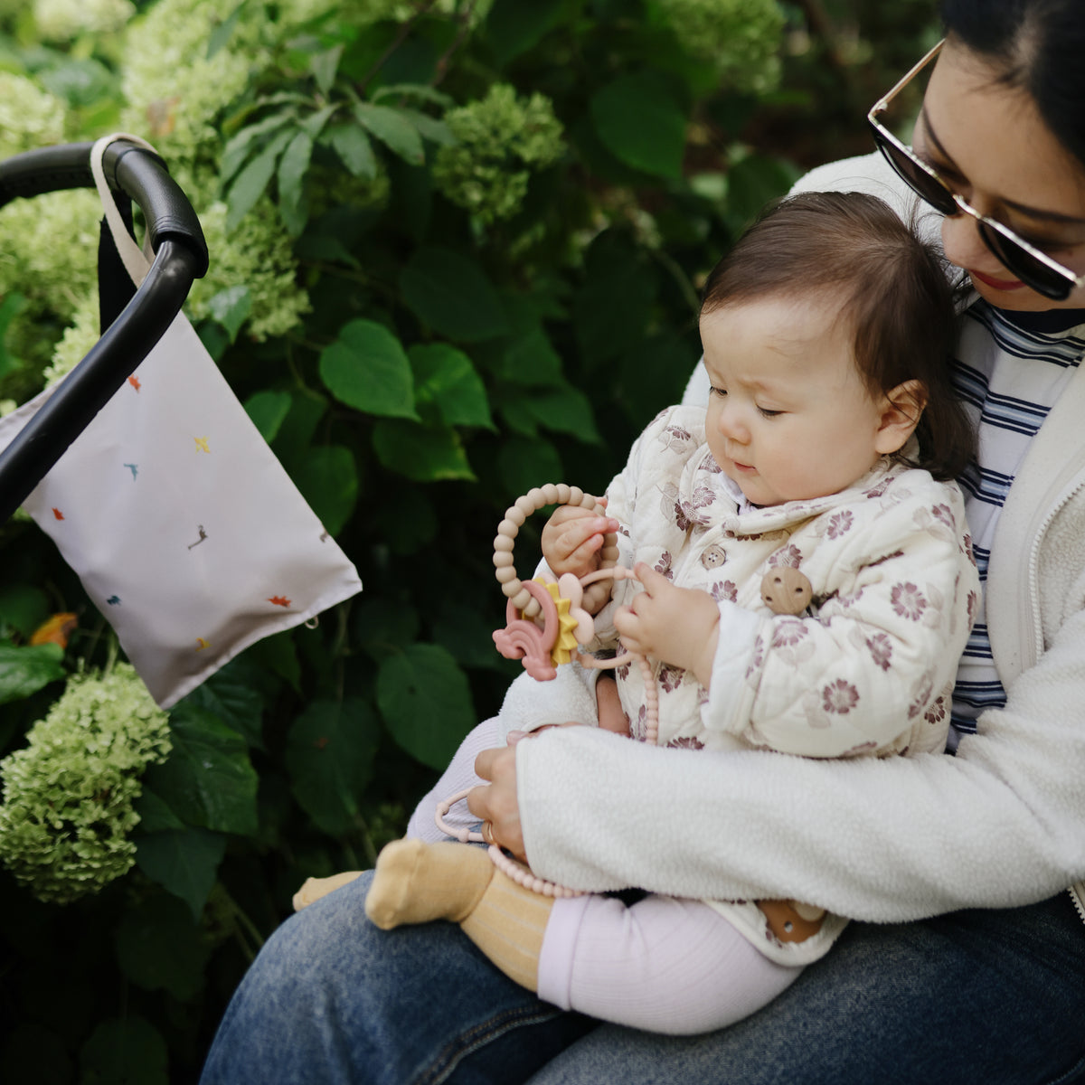 A baby outdoors with a woman, holding mushie Adjustable Silicone Strap, amid green plants.