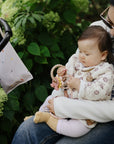 A baby outdoors with a woman, holding mushie Adjustable Silicone Strap, amid green plants.