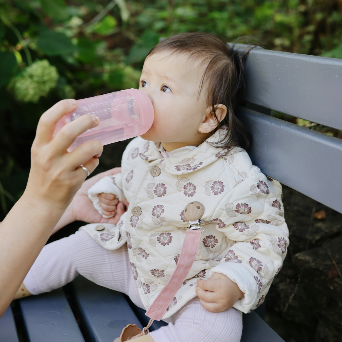 Baby on a bench, fed by an adult with a mushie Soft Spout Sippy Cup, wearing a floral jacket.