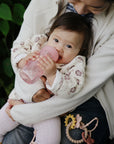 Baby with mushie Soft Spout Sippy Cup on lap, pink, silicone, toys beside.
