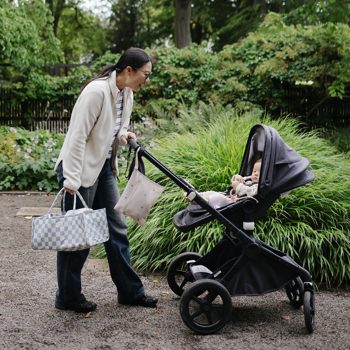 Woman with mushie Diaper Caddy, pushing stroller amid greenery.