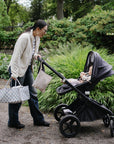 Woman with mushie Diaper Caddy, pushing stroller amid greenery.