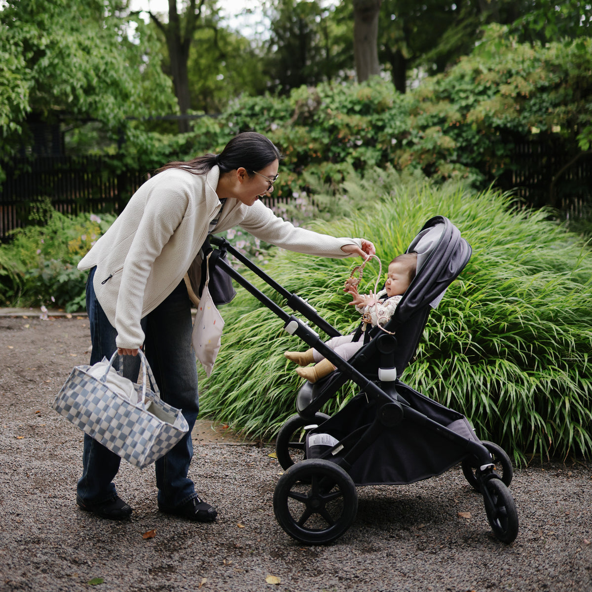 Woman hands baby a mushie Adjustable Silicone Strap, both smiling together outdoors in the park.