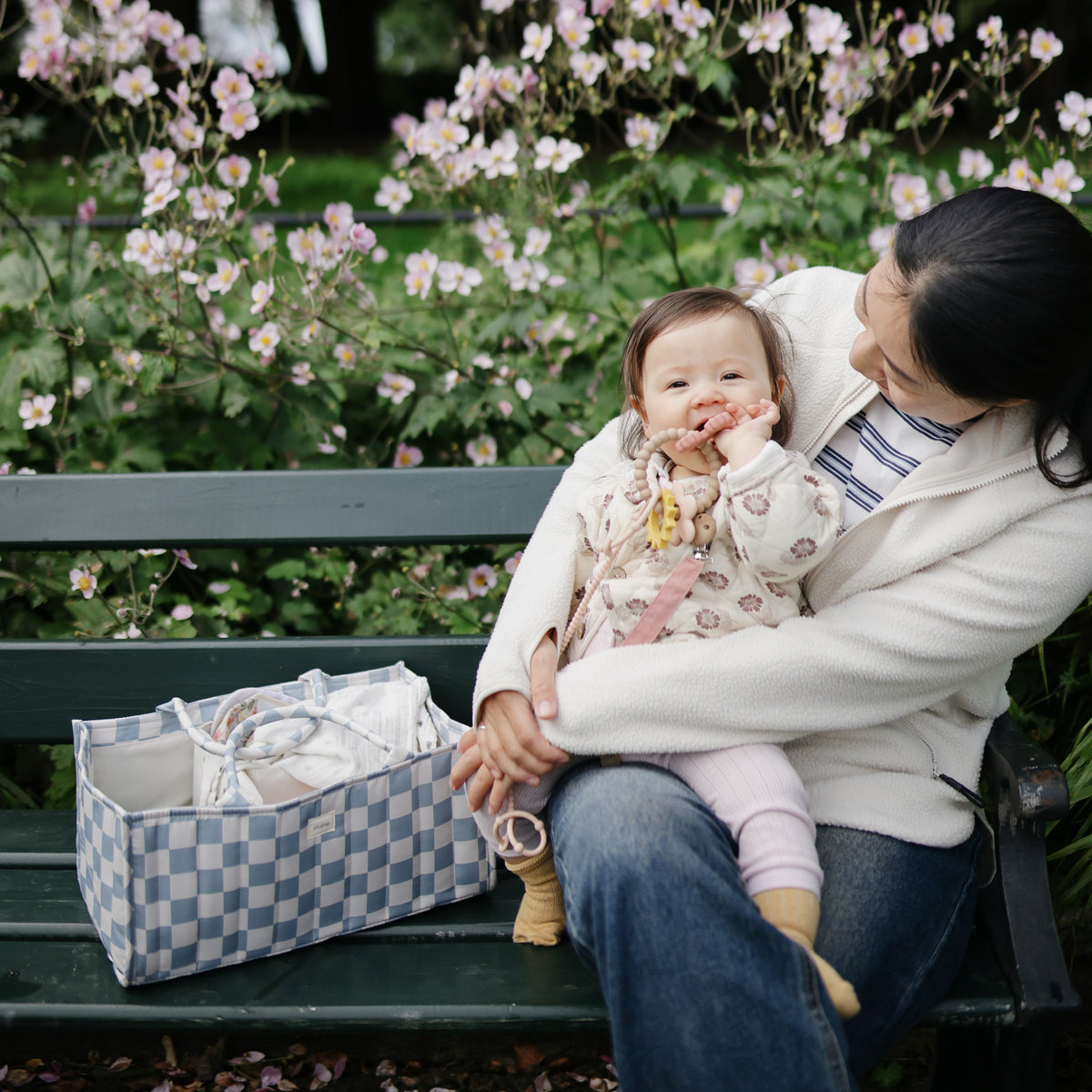 Woman with baby on bench near mushie Diaper Caddy; pink flowers bloom in the background.