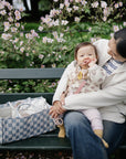 Woman with baby on bench near mushie Diaper Caddy; pink flowers bloom in the background.