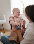 Smiling baby in mushie Bear Poncho Towel, holding a toy on woman’s lap indoors.