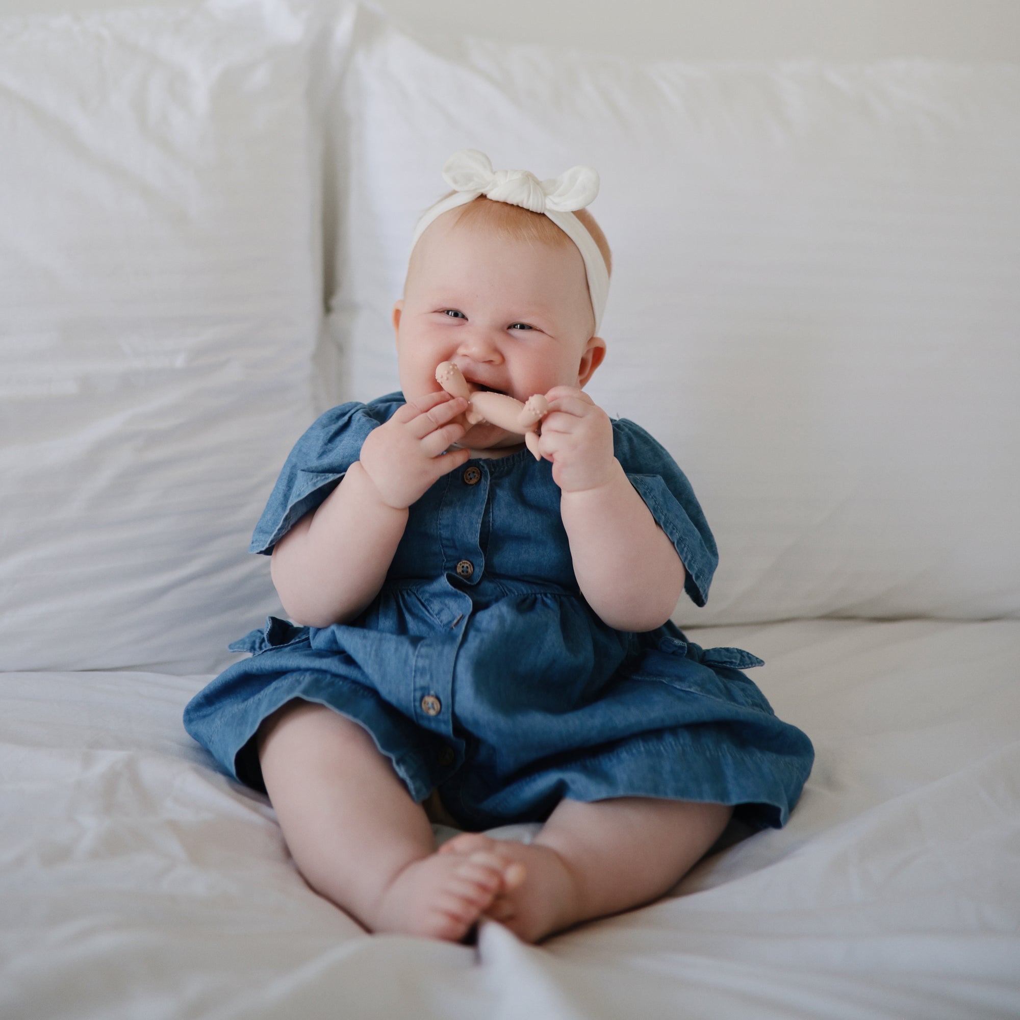 Smiling baby in blue dress holding mushie Figurine Teether while sitting on a bed.