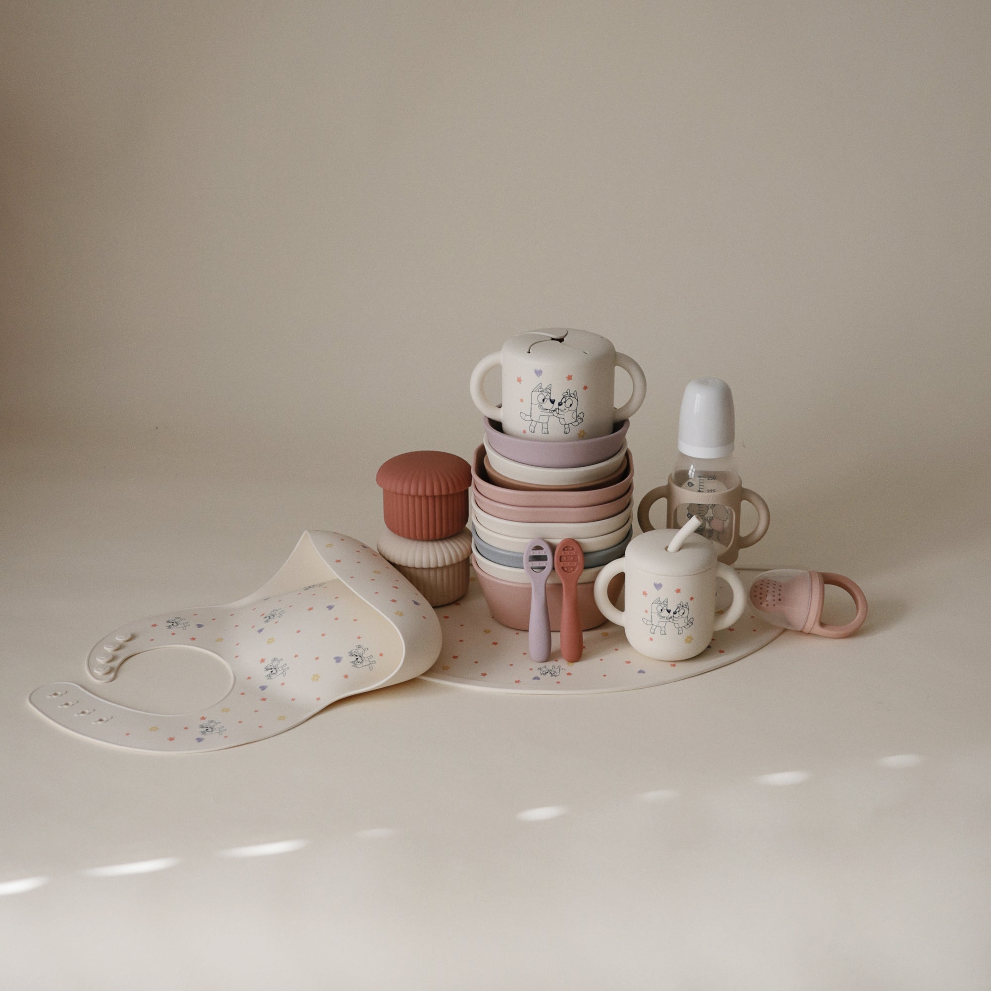Mushie Bluey Gift Set: neutral silicone cups, plates, utensils, bib &amp; bottle on a table.