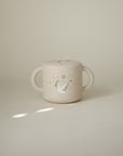 Mushie Snack Cup in beige, spill-proof with two handles and a cute bird illustration for toddlers.