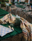 A sippy cup, mushie Organic Cotton Muslin Swaddle Blanket, and bag on a green outdoor table.