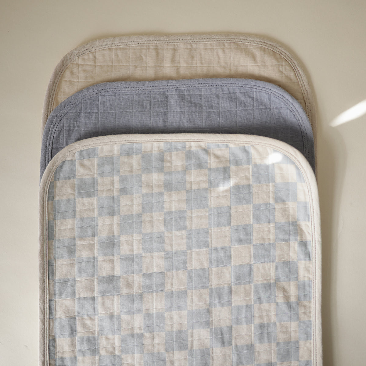 Three Mushie Changing Pad Liners in checkered, solid blue, and solid tan patterns.