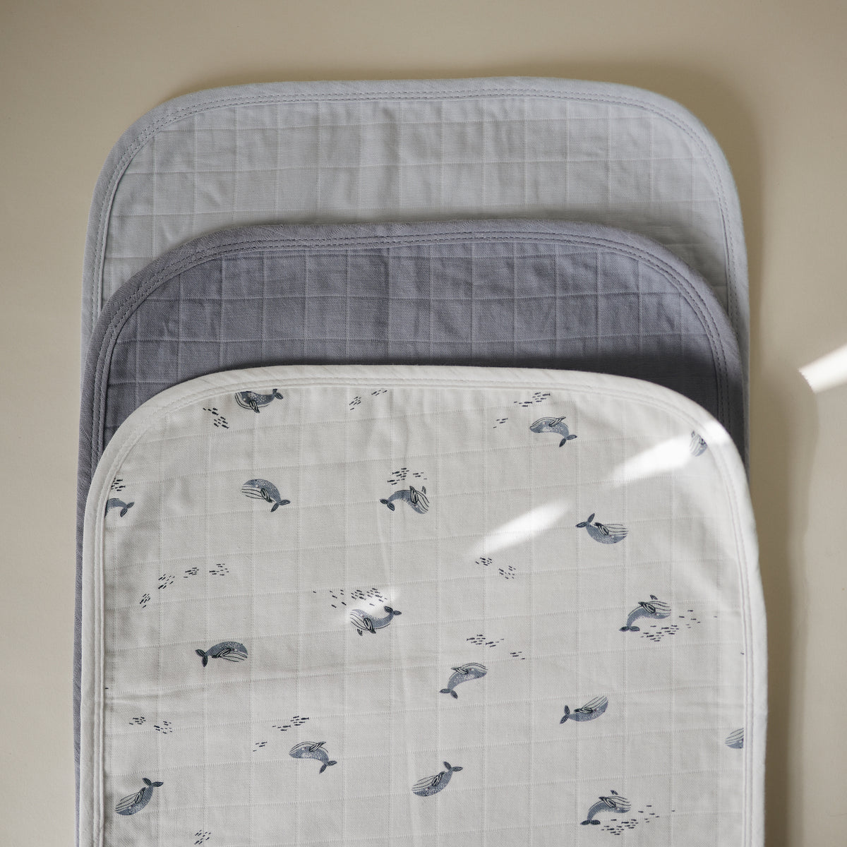Three stacked Mushie Changing Pad Liners in white with whale patterns, grey, and light blue.