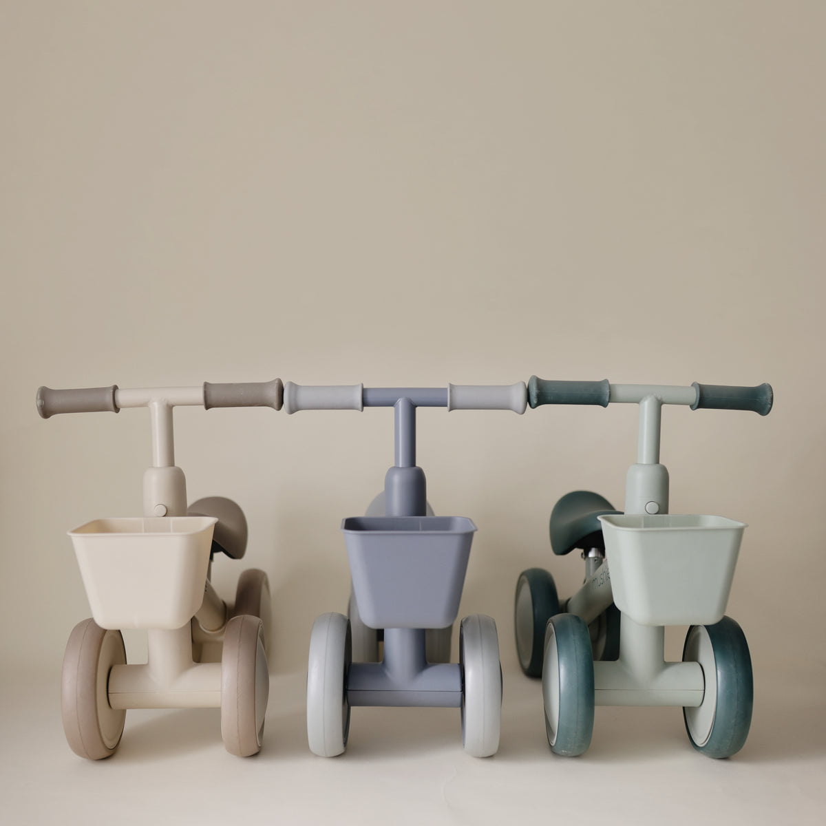 Three mushie Mini Toddler Bikes in beige, blue-grey, and sage green, each with a front basket.