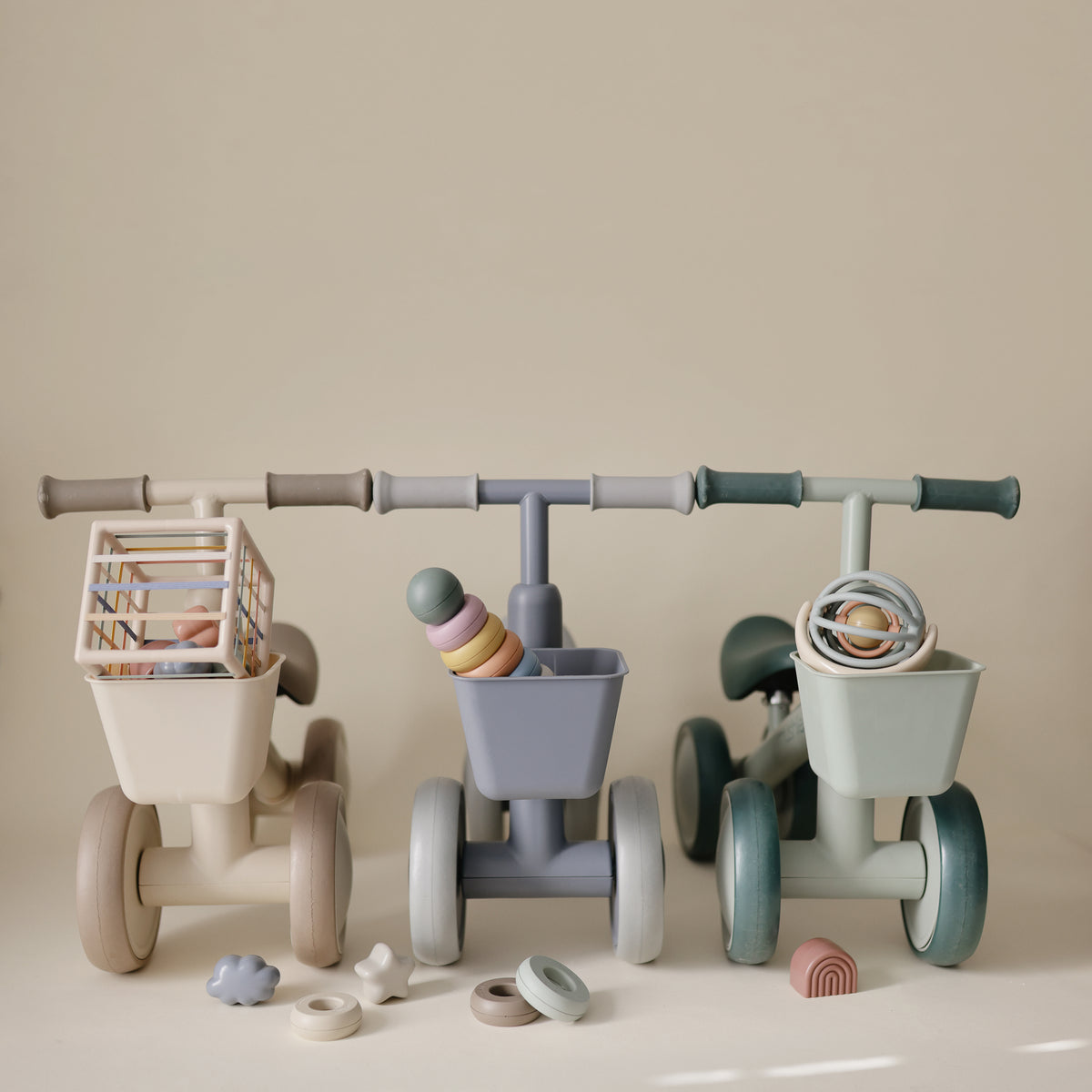 Three beige, blue, and sage green mushie Mini Toddler bikes, each with a front basket and toy.