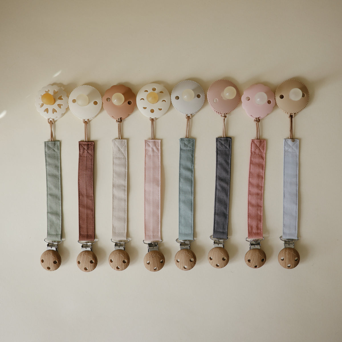 Mushie Linen Pacifier Clip 2-Pack, pastel colors, wooden clips on beige background.