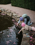 Two children play by a pond with mushie Bath Boats, wearing pastel hats and jackets.