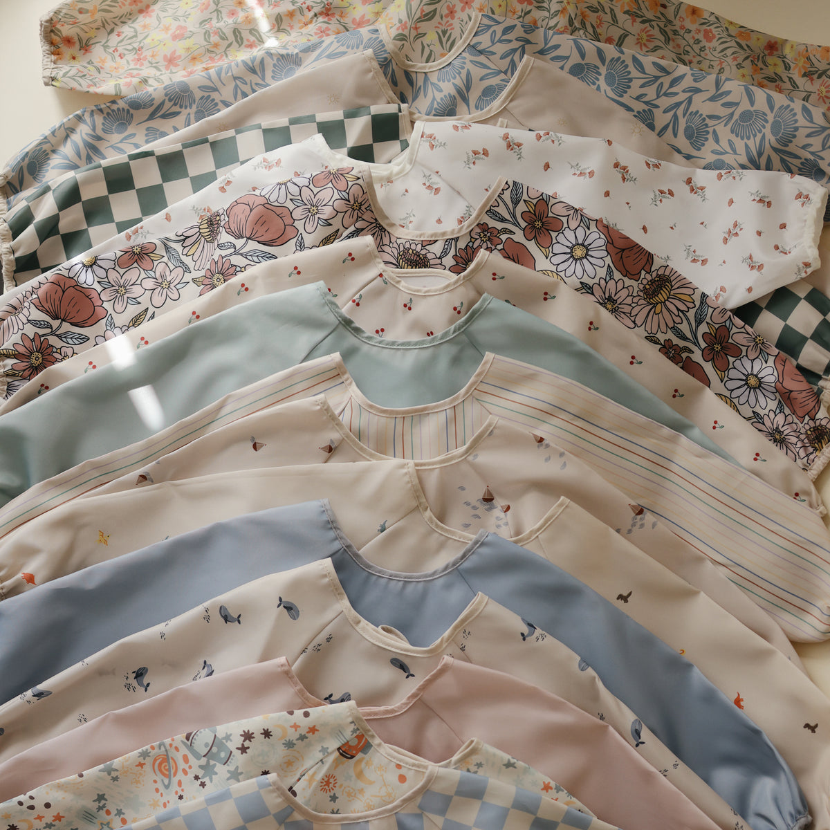 A stack of patterned, water-resistant mushie Long Sleeve Bibs with floral and checkered designs.