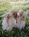 Two young children in white dresses and wings explore a field of white blooms. One child, focused on baby’s comfort, holds a FRIGG Butterfly Anatomical Silicone Pacifier 2-Pack (0-6 Months) by FRIGG like a precious treasure.