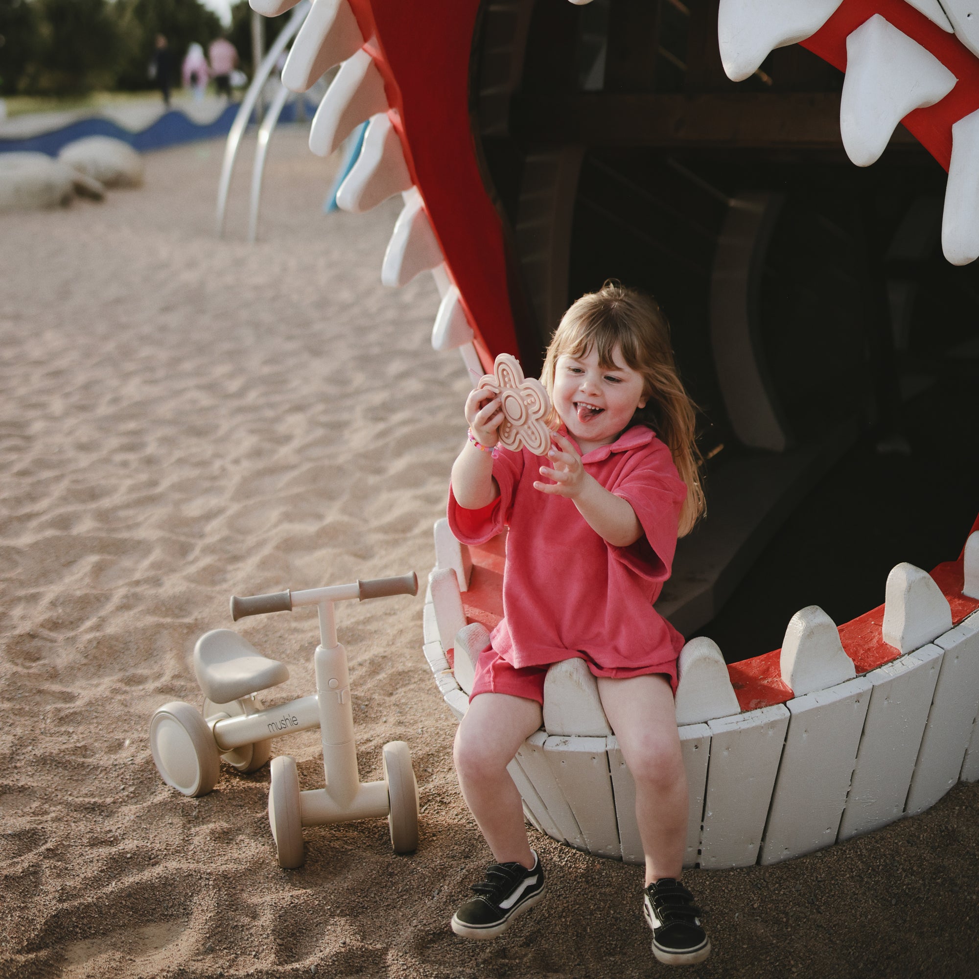 A girl smiles on a playground, holding the mushie Silicone Sensory Toy in her mouth.