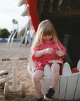 Young girl plays with mushie Silicone Sensory Toy, sitting near a white wooden structure.