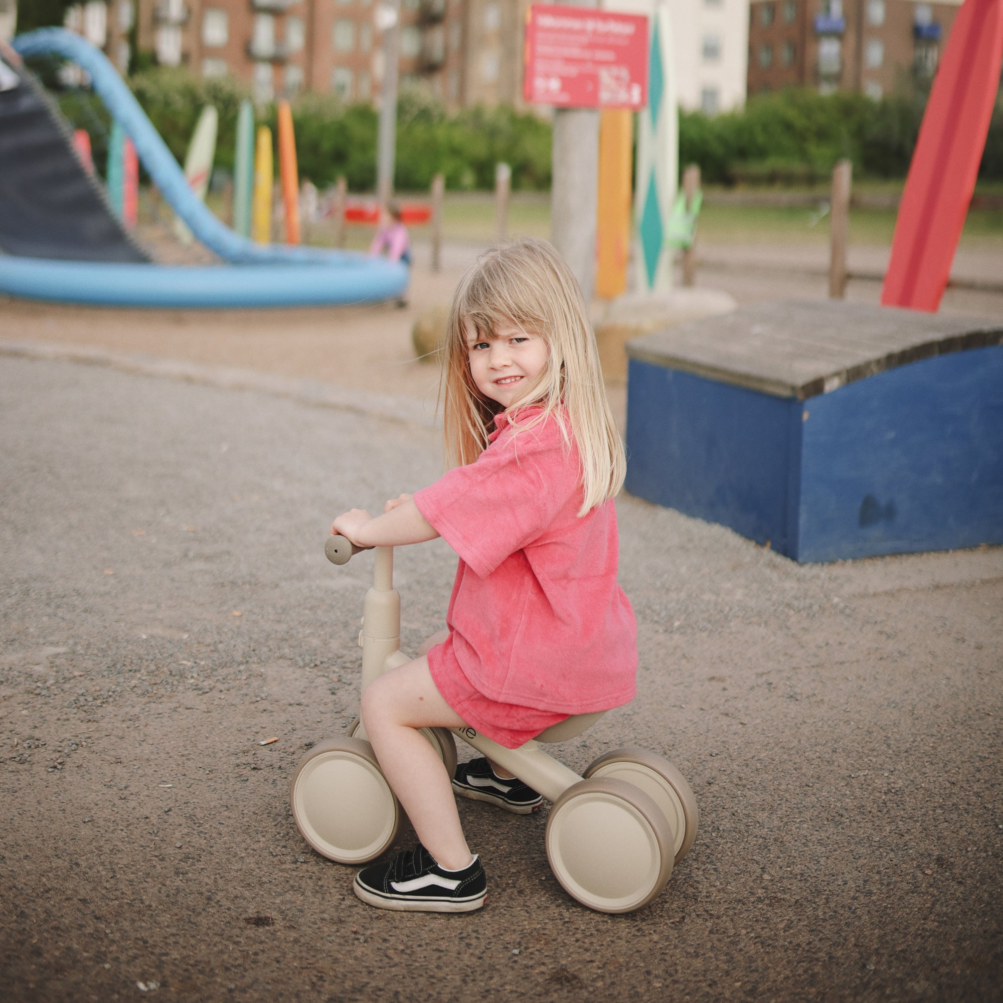 Young girl in pink rides a beige mushie Mini Toddler Bike at the playground, smiling happily.
