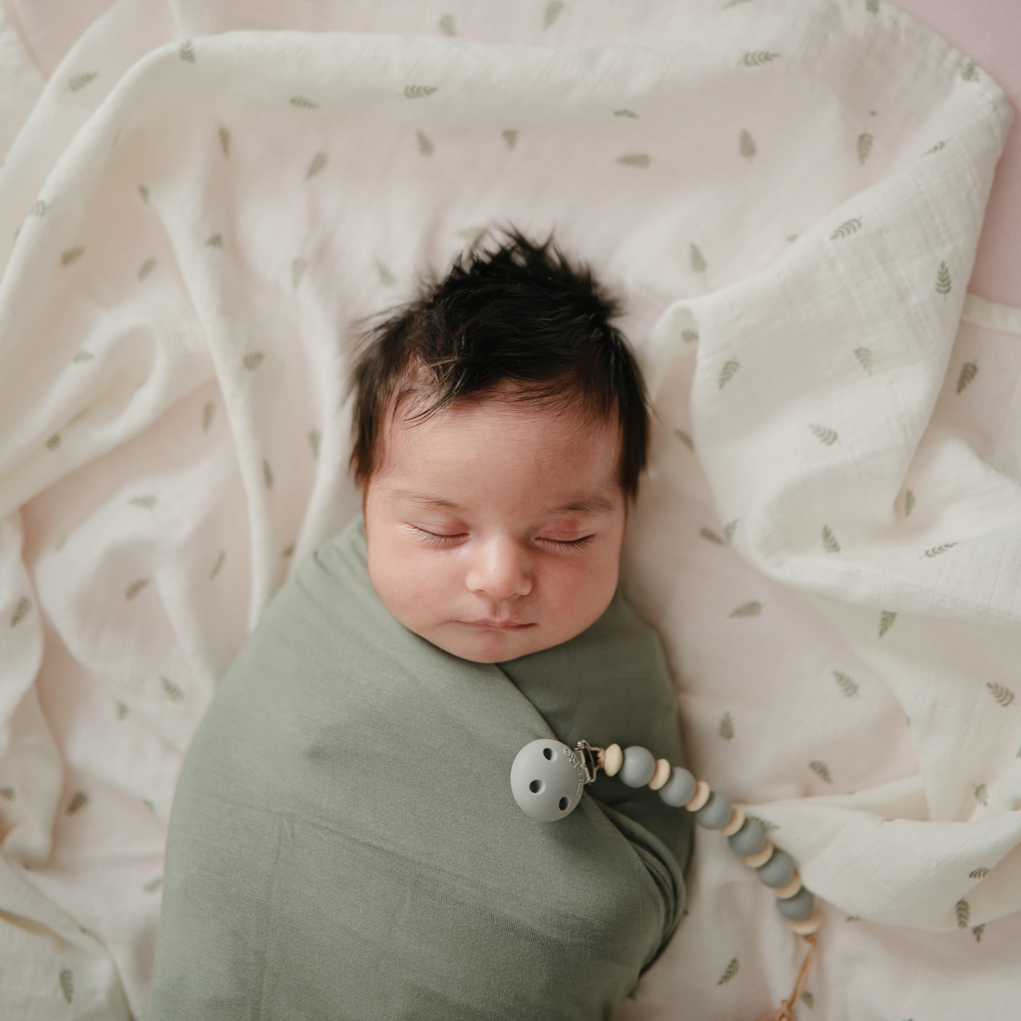Baby with dark hair wrapped in mushie Organic Cotton Muslin Swaddle Blanket, on white cloth.