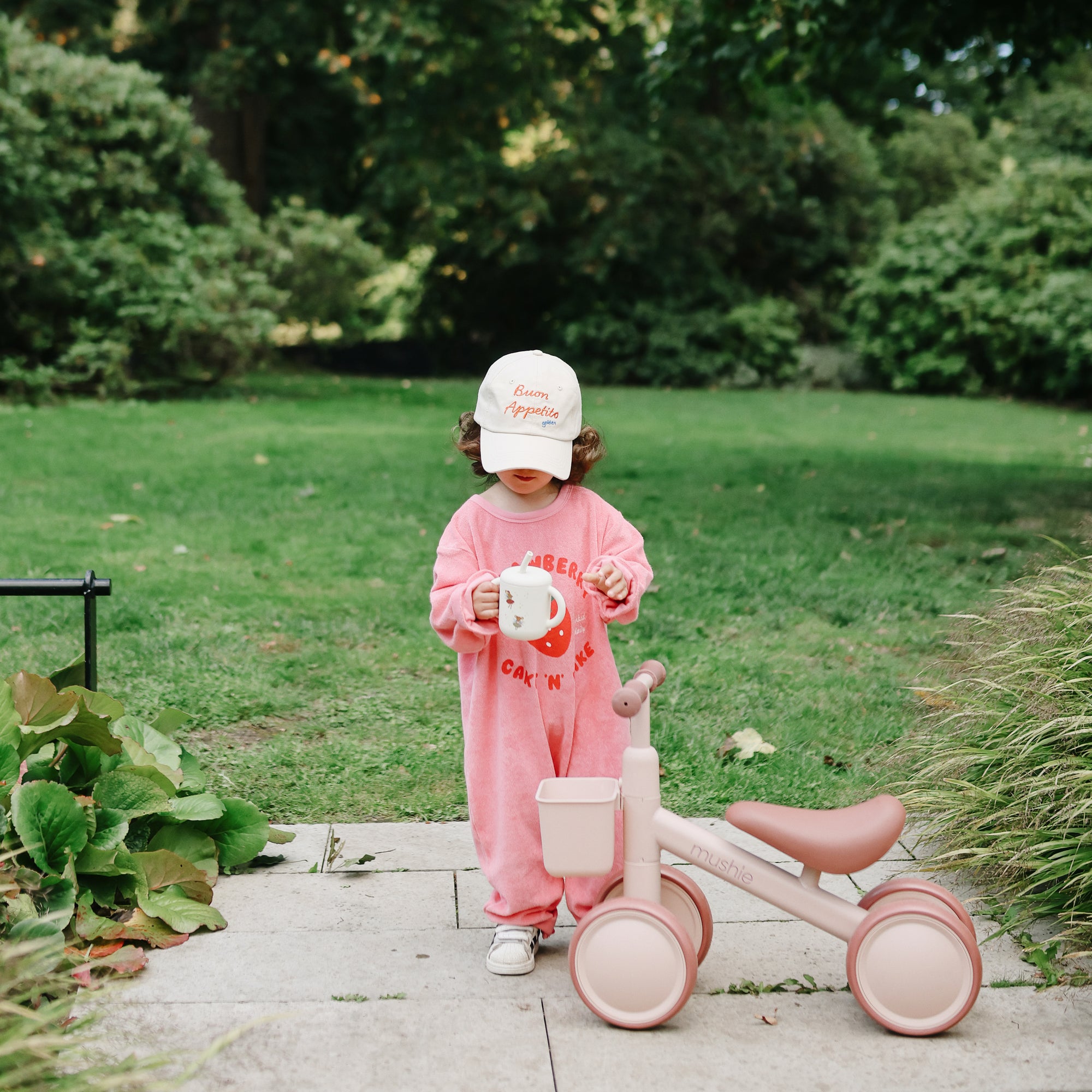 Toddler with Mushie Silicone Training Cup + Straw stands by a pink tricycle in the park.