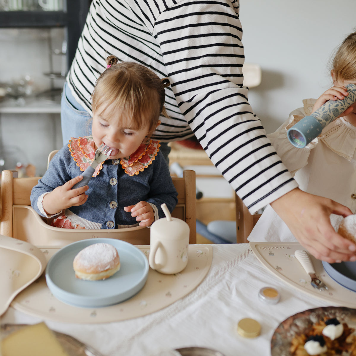 Toddler eats pastry from Mushie Classic Silicone Suction Plate; adult helps another child nearby.
