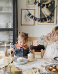 Two kids at Hanukkah; one eats, one uses a Mushie Classic Silicone Suction Plate.