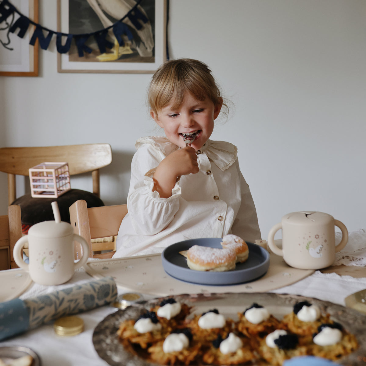 Happy child enjoys a donut on a Mushie Classic Silicone Suction Plate, snacks and drinks close by.