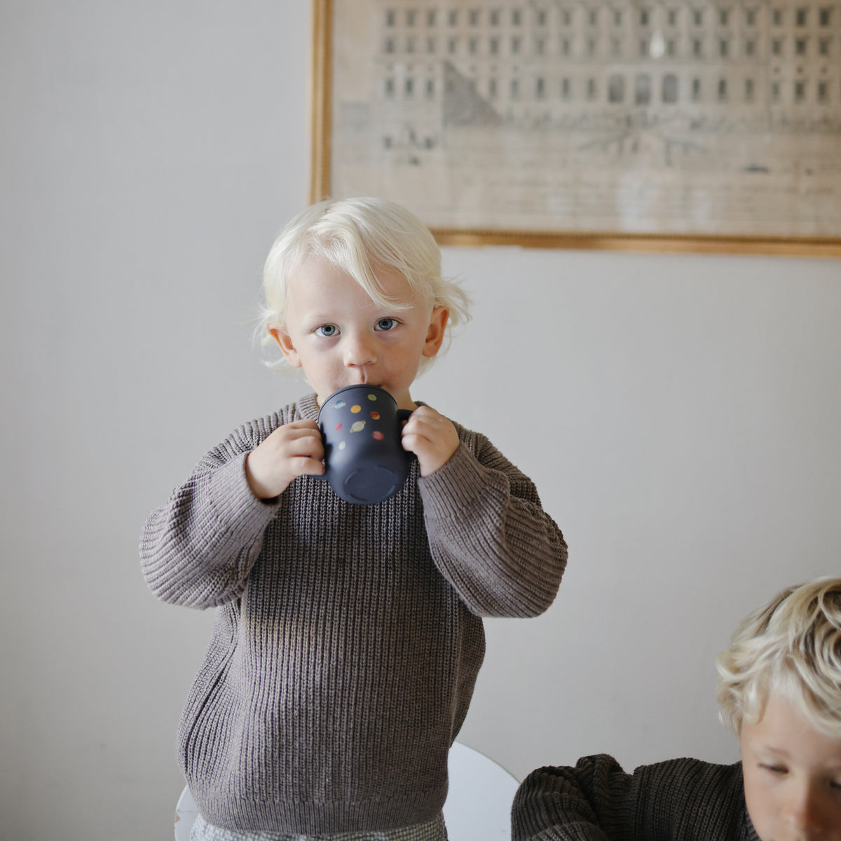 Blonde toddler in brown sweater drinks from Mushie Silicone Training Cup + Straw indoors.