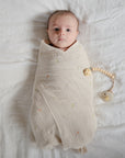 A baby wrapped in a mushie Organic Cotton Muslin Swaddle Blanket lies beside a pacifier clip.