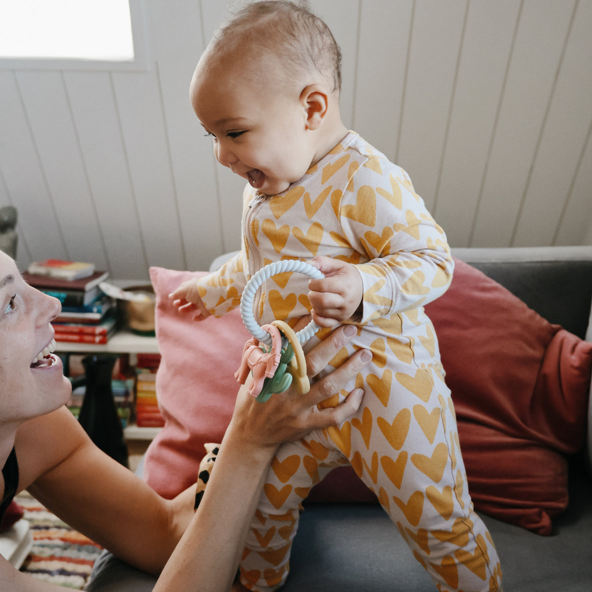 Baby in heart PJs holds mushie Teething Gift Set while being lifted by an adult on the couch.