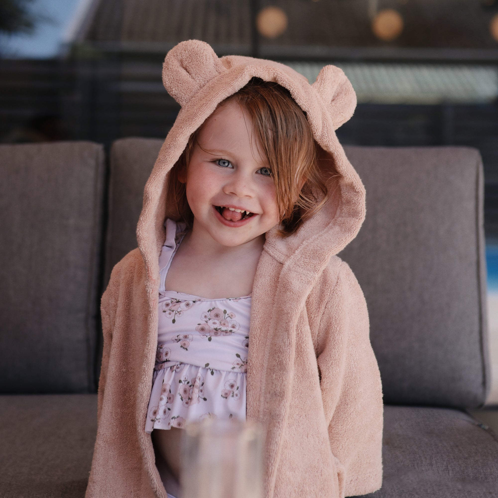 Smiling child in mushie's Bear Robe over a floral swimsuit, sitting on a couch.