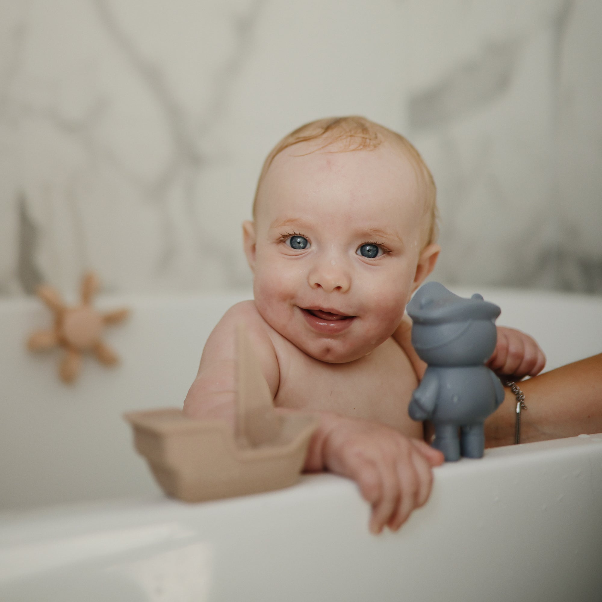 Smiling baby in a tub with mushie Mold Free Bath Play Set and floating toys, easy to clean.