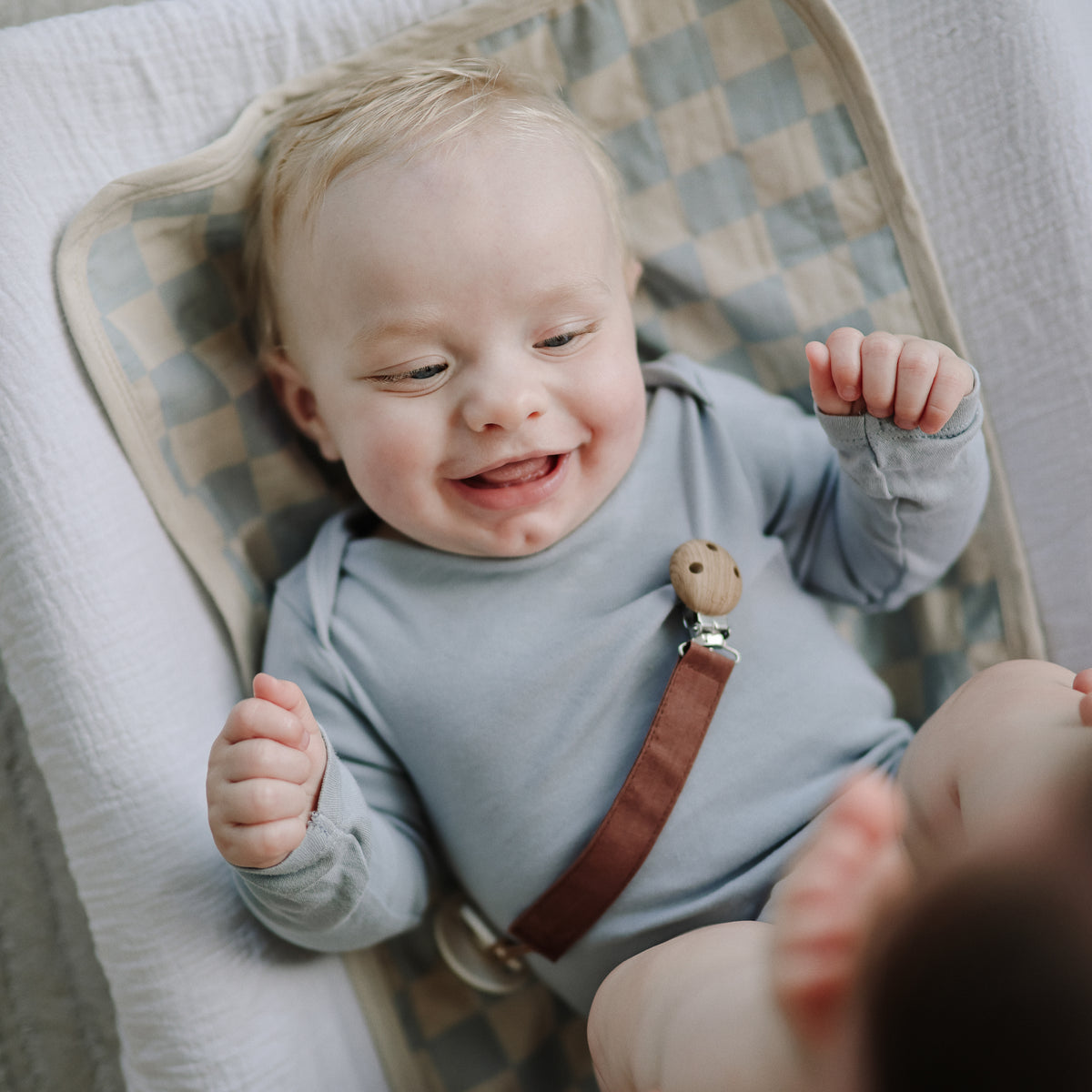 Smiling baby in a gray onesie holds mushie’s Linen Pacifier Clip 2-Pack while lying on a mat.