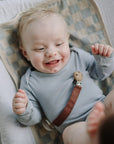 Smiling baby in a gray onesie holds mushie’s Linen Pacifier Clip 2-Pack while lying on a mat.