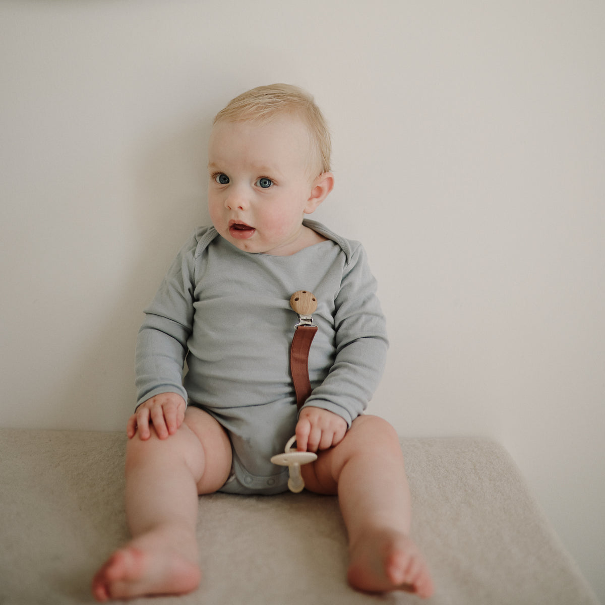 Baby holds a mushie Linen Pacifier Clip 2-Pack, wearing gray onesie, looking to the side.