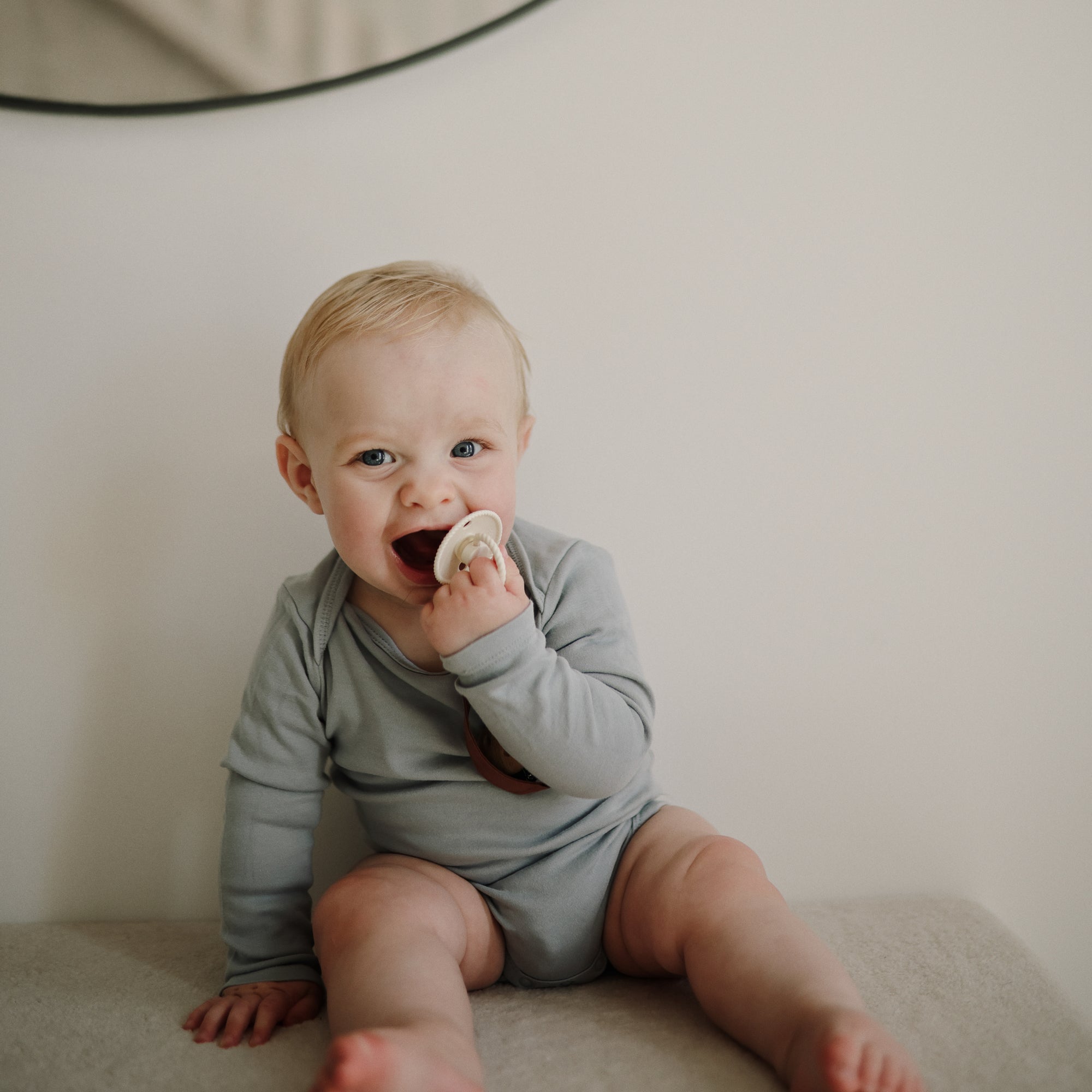 Baby in a grey onesie holds a FRIGG Rope Silicone Pacifier 2-Pack and smiles slightly.