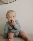 Baby in a grey onesie holds a FRIGG Rope Silicone Pacifier 2-Pack and smiles slightly.