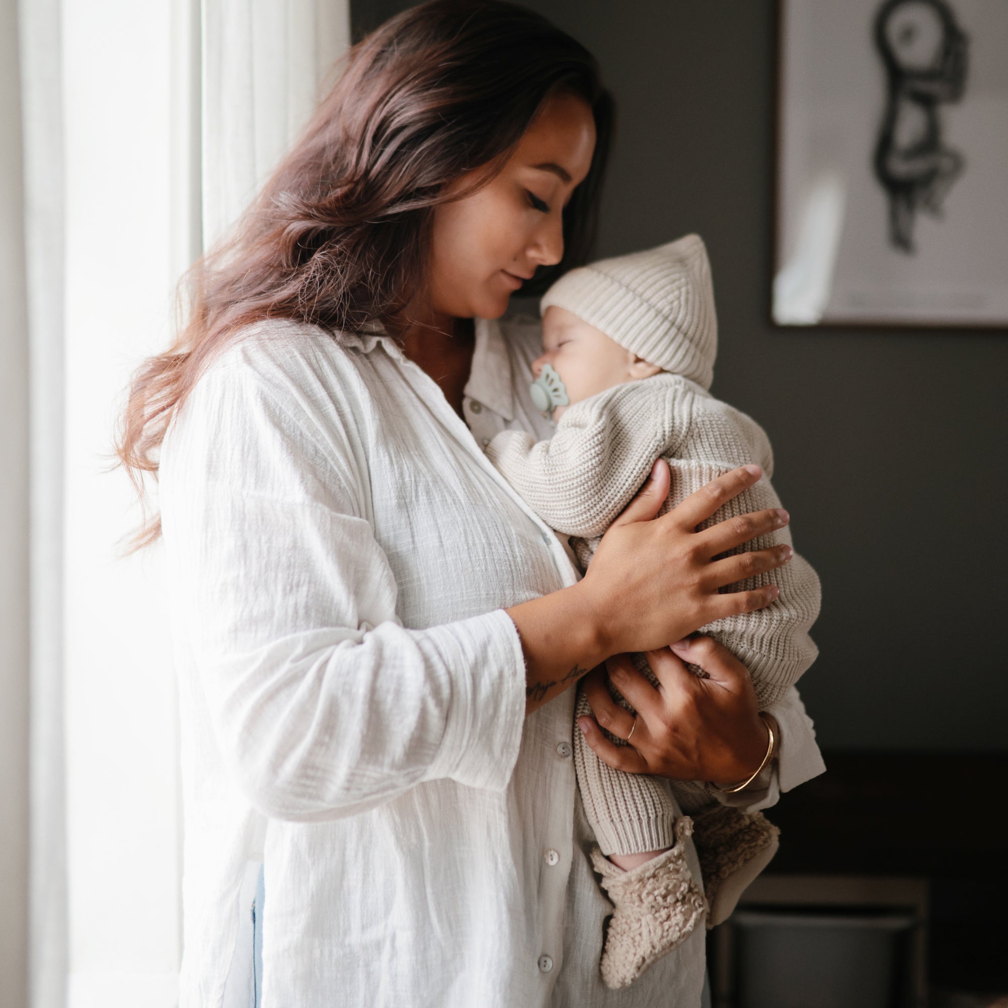 A woman in a white shirt gently holds a sleeping baby in beige knitwear near a bright window, soothing the infant with a FRIGG Butterfly Anatomical Silicone Pacifier 2-Pack (0-6 Months) by FRIGG.