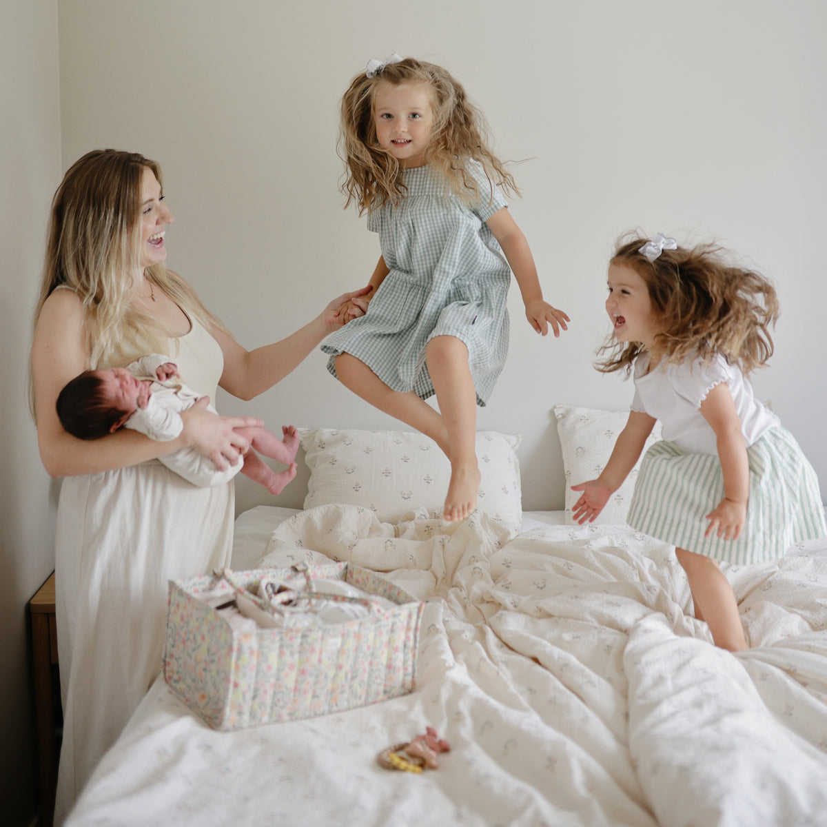 Woman holds baby as two girls jump on bed near a mushie Diaper Caddy.