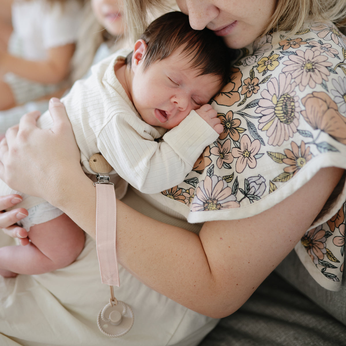 Newborn in arms, mushie Linen Pacifier Clip 2-Pack with silicone clasp visible nearby.