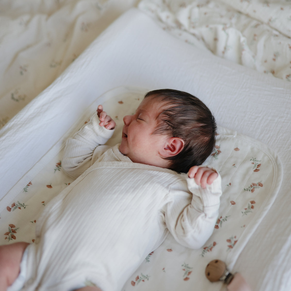 Newborn in white onesie on mushie Extra Soft Muslin Changing Pad Cover, hand raised near face.
