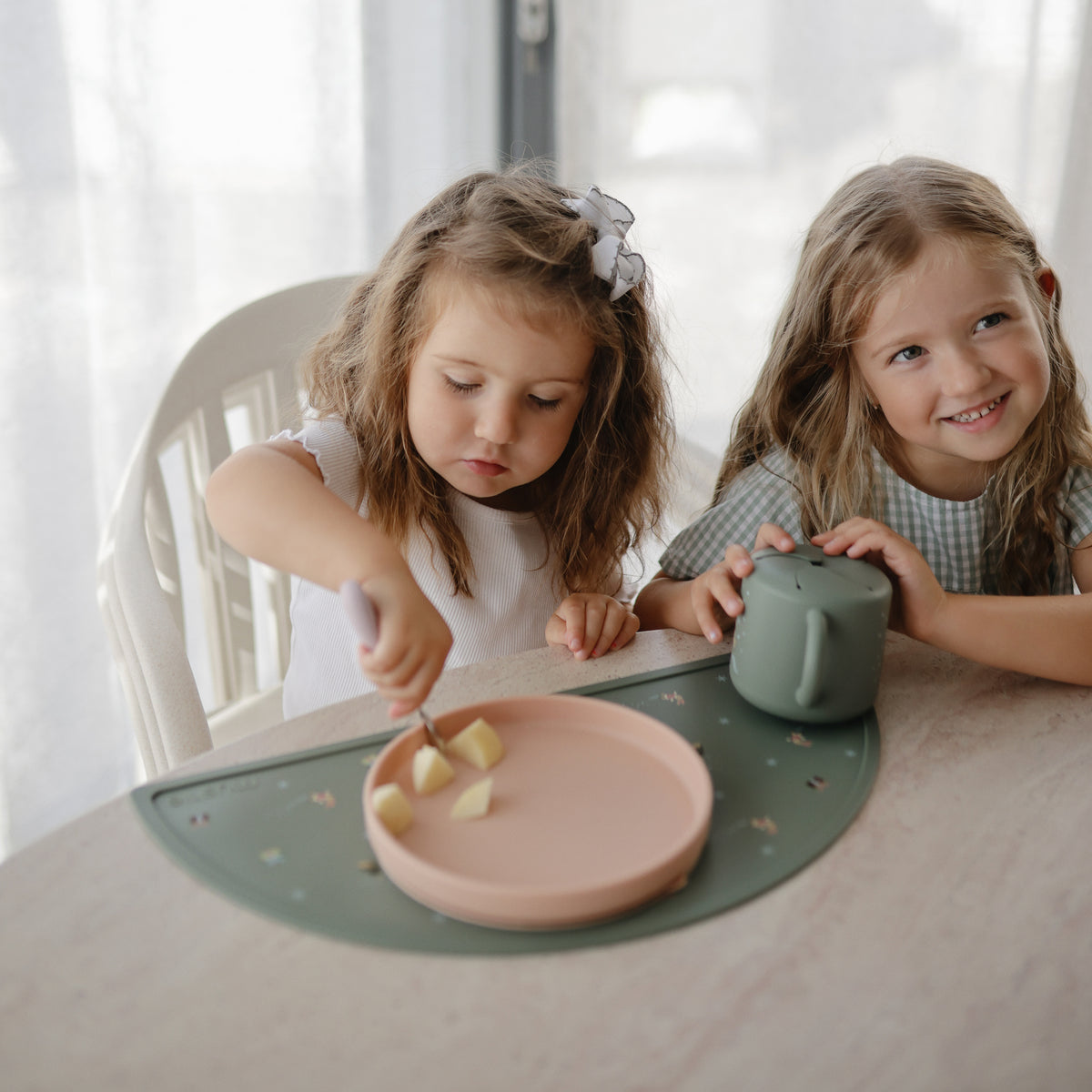Two young girls sit at a table; one uses a Mushie Classic Silicone Suction Plate for snacks.