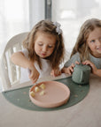 Two young girls sit at a table; one uses a Mushie Classic Silicone Suction Plate for snacks.