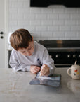A boy writes at a counter with a Mushie Silicone Training Cup + Straw and a mug nearby.