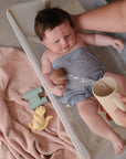An adult gently bathes a baby in a small plastic tub by pouring water with the Mushie Bath Rinse Cup. The baby, in gray cloth, is holding a toy and surrounded by a pink towel, yellow squirrel toy, and green binoculars.