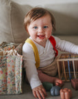 Lifestyle image of a baby sitting on a couch with the Elastic Shape Sorting toy and a Pastel Blooms Diaper Caddy around, wearing a white shirt, yellow suspenders, and a red bib