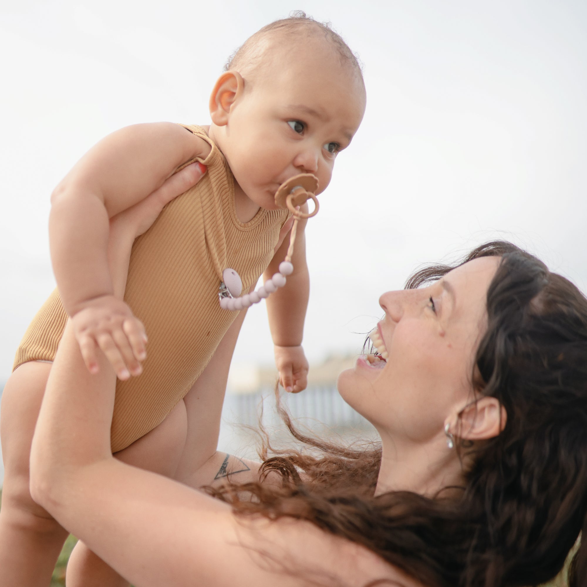 A woman with long brown hair smiles up at a baby in a tan onesie using the FRIGG Daisy Natural Rubber Pacifier (Limited Edition), holding the baby up outdoors on a bright, sunny day.